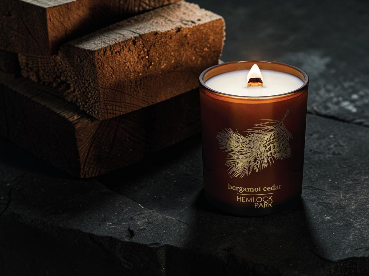 Hemlock Park Bergamot Cedar Apothecary Candle with a glowing wood wick flame on dark slate stone beside stacked cedar logs